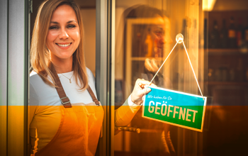 VisitorApp Business Eine glückliche Ladeneigentümerin dreht das Türschild an der Eingangstüre um und zeigt damit, dass der Laden geöffnet hat.